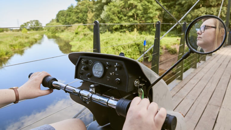 The image shows a person riding a mobility scooter across a wooden suspension bridge over a river. The scene features lush green trees and bushes along the riverbank, reflecting in the calm water. The scooter's control panel and rear-view mirror are visible, with the person’s face reflected in the mirror. The rider is gripping the handlebars, and the focus is on the scooter's dashboard with various buttons and a display. The atmosphere is serene, with a clear, sunny day and natural surroundings.